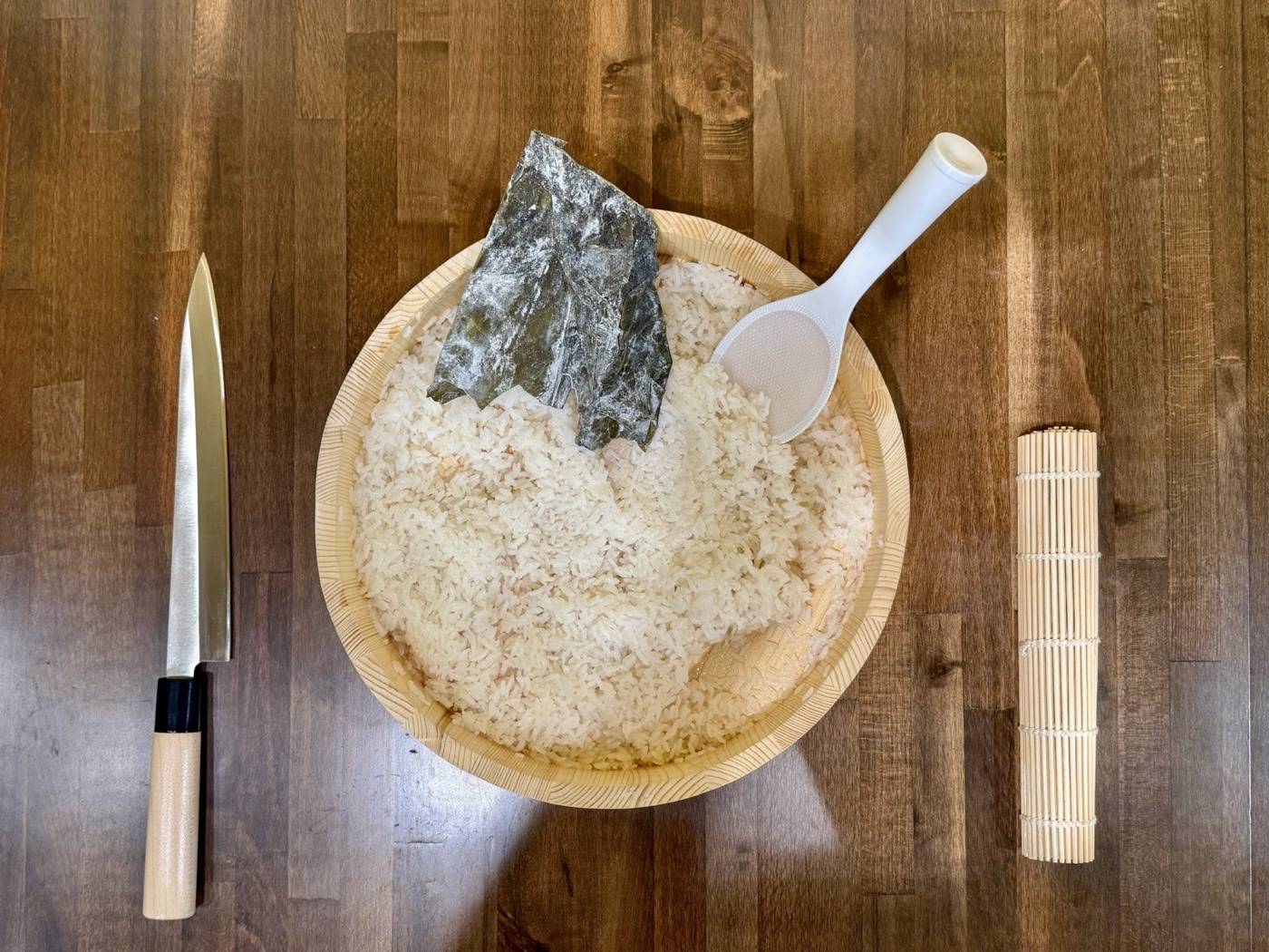 Seasoned rice in a hangiri with nori and a rice paddle, a yanagiba to one side and a rolling mat to the other, on a wooden counter